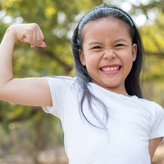 A young girl wearing a white t-shirt and showing off her muscles smiles wide after finishing a dentist’s appointment in Fort Worth